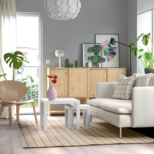 Sala de estar moderna con alfombra de rayas rojas, sofá gris, plantas y balcón abierto.