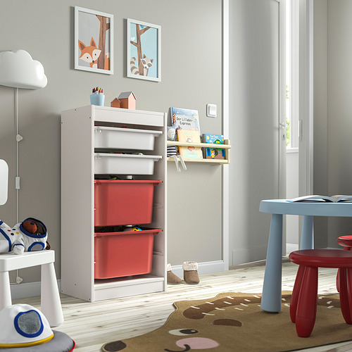 Una unidad de almacenamiento TROFAST con marco blanco y cajas rojas, colocada en una sala de juegos para niños. Tiene capacidad para juguetes y libros, y está al lado de una pequeña mesa y taburetes.