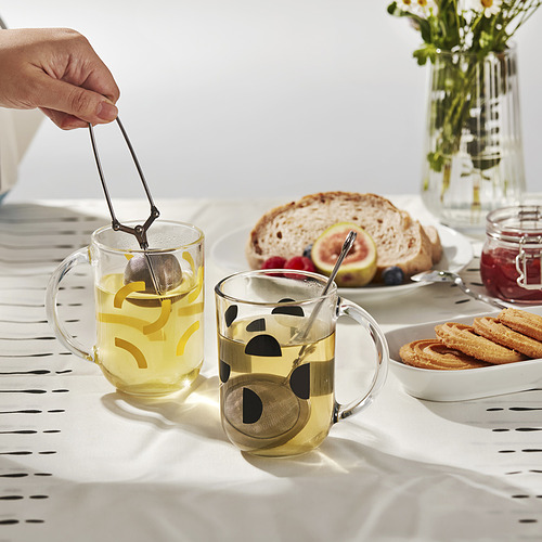 La mano sostiene el colador de té en una taza de vidrio. Las tazas tienen granos de café, patrones de hojas. Mesa con pan, bollería, mermelada, flores.