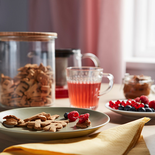 Una mesa de desayuno con galletas de letras KAFFEREP en un plato verde, bayas frescas y una taza de vidrio de té rojo, colocada sobre un paño amarillo.