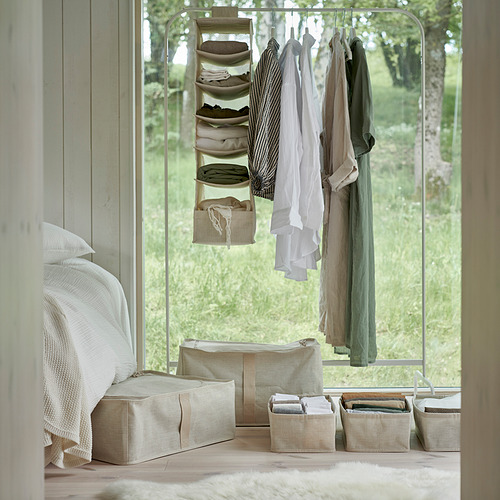 Dormitorio con organizador de tela para colgar, beige, compacto, sosteniendo ropa doblada, junto a un espejo grande, debajo del perchero, sábanas blancas sobre la cama.