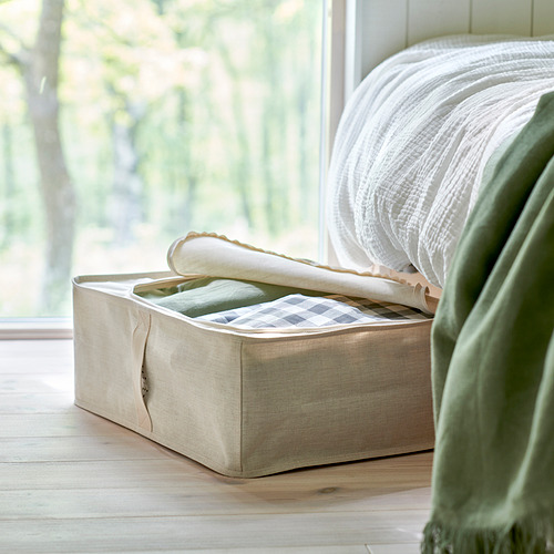 Caja de almacenamiento de tela beige junto a la cama, guarda la ropa, manta verde cerca, protege del polvo.