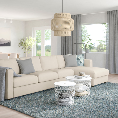 Salón moderno con sofá en forma de L color crema, mesitas de centro, lámpara colgante, alfombra y cortinas grises, luz natural.