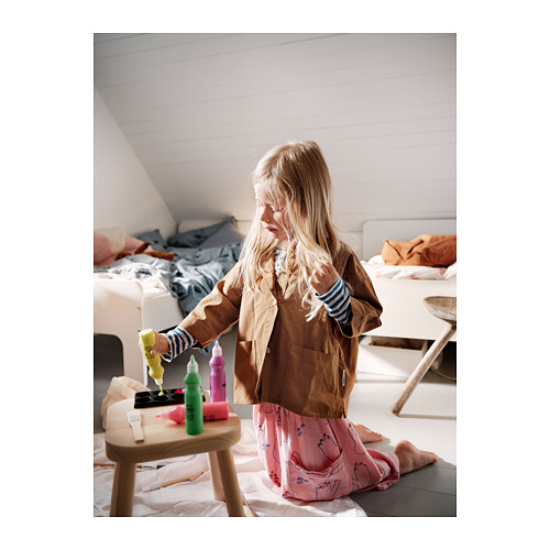 Una niña está haciendo manualidades con el set de arte MÅLA con pinceles, tijeras y rotuladores en un taburete de madera en una habitación con camas.