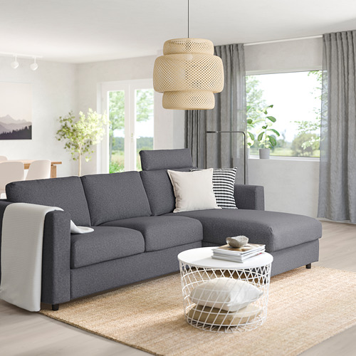 Sala de estar moderna con sofá VIMLE gris, tumbona, almohadas. Mesa de centro con libros, gran ventanal, suelo de madera, cortinas grises, lámpara colgante.