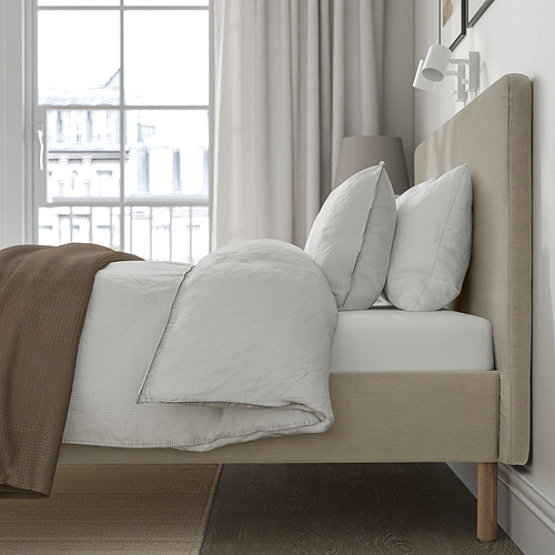Cama con cabecera suave y acolchada, sábanas blancas, manta marrón y gran ventana.