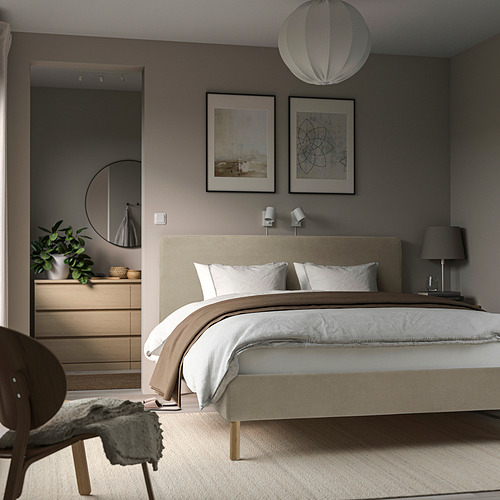Dormitorio moderno con cama beige, cabecero de felpa, ropa de cama blanca, paredes grises, luz redonda, arte, tocador, espejo, lámpara negra.