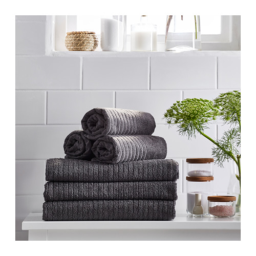 La imagen muestra toallas VÅGSJÖN de color gris oscuro enrolladas y dobladas, incluidas toallas de mano y de baño, expuestas en estantes blancos con una planta verde.