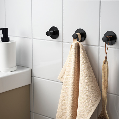 Tres ganchos negros en la pared de azulejos, dos que sostienen toallas y un cucharón de madera que cuelga. Fácil de instalar con cinta adhesiva.