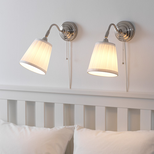 Dos lámparas de pared con pantallas beige orientadas hacia abajo iluminan una cama blanca con almohadas. Tire de los cables para los interruptores visibles.
