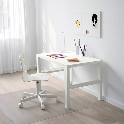 Un pequeño escritorio blanco ajustable con ruedas, una silla y coloridos útiles escolares, colocado debajo de una ventana con cortinas ligeras.