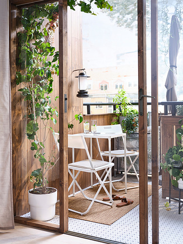Pequeño y acogedor balcón con mesa y sillas de metal blanco, plantas verdes y alfombra natural, visto desde el interior.