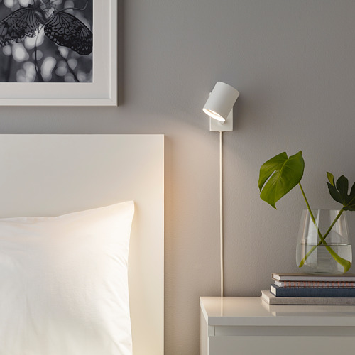 Dormitorio minimalista con lámpara SOLHETTA que ilumina plantas y libros, paredes grises y estampado de mariposas sobre la cama.