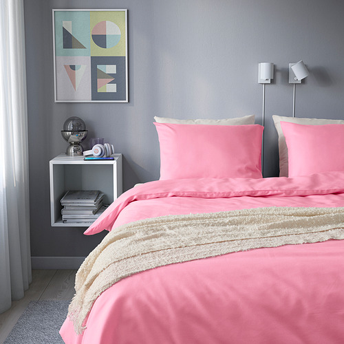 Edredón PILTANDVINGE de color rosa vibrante colocado en una cama acogedora, manta blanca, mesita de noche con libros, obras de arte coloridas arriba.