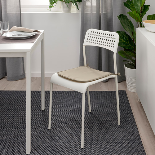 Escena de comedor moderno con almohadilla de silla BRÄMÖN blanca, beige, en silla blanca cerca de la mesa con platos y vidrio, contra la ventana y las plantas.