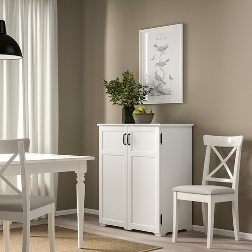 Mueble GREÅKER blanco con 4 puertas, cuadro y planta entre la ventana y la mesa de comedor, con una silla al lado.