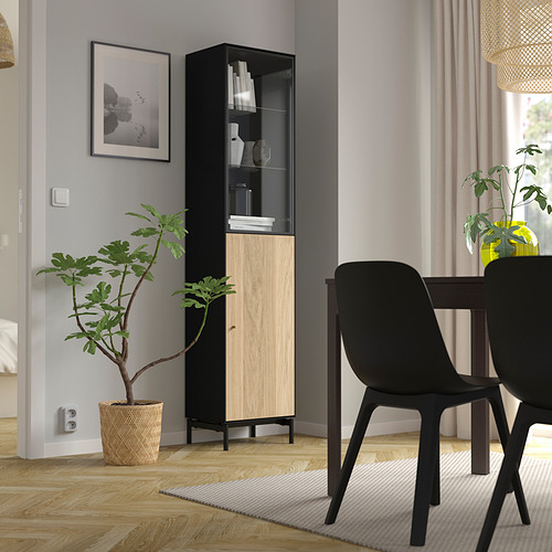 Mueble BOASTAD negro y roble con puerta de cristal que exhibe libros y jarrones, flanqueado por una alta planta verde y sillas de comedor negras, contra una pared gris.