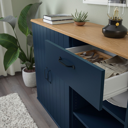 Armario azul con cajón abierto que deja ver cubiertos y paños; Tapa de madera con plantas, libros y lámpara.