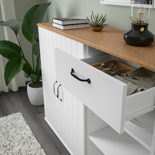 Mueble blanco con cajón abierto que deja ver platos y paños, tapa de madera con libros y planta.