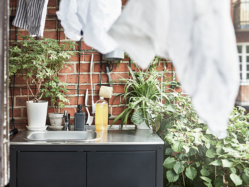 Pequeño huerto urbano con plantas en macetas, fregadero, toallas y herramientas dispuestas sobre una pared de ladrillo, reflejando un ambiente de cocina exterior acogedor y práctico.