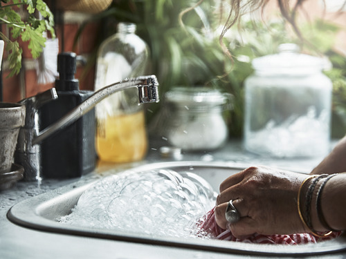 Persona enjuagando verduras en un fregadero de plata con agua corriente, rodeada de botellas y vegetación.