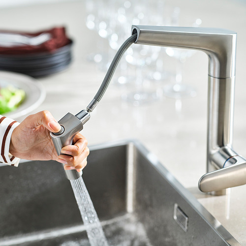 Toma a mano el rociador extraíble de los grifos MAJSJÖN , el agua fluye desde él hacia el fregadero de la cocina de acero inoxidable.