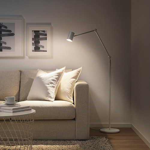 Una lámpara de pie negra de NYMÅNE junto a un sofá beige, que añade una iluminación moderna a un acogedor rincón de la sala de estar.