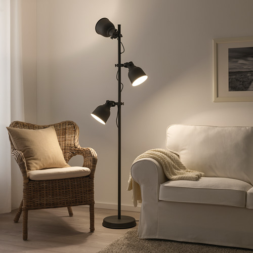 Lámpara de pie HEKTAR, negra, diseño industrial, grandes pantallas metálicas, altura regulable, se alza junto a silla de mimbre y sofá beige.