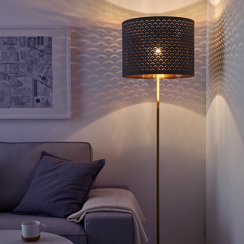 Una sala de estar moderna con una lámpara de pie NYMÖ que proyecta una luz estampada en la pared. La lámpara tiene una base de color latón y una pantalla perforada en color negro.