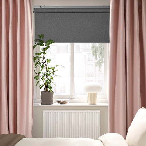 Ventana del dormitorio con persiana enrollable gris, cortinas de textura lineal de color rosa suave, lámpara blanca y planta verde alta en el alféizar, radiador blanco debajo.