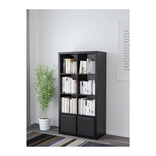 Estantería KALLAX negra de pie, llena de libros y objetos de decoración, con una planta en maceta al lado.