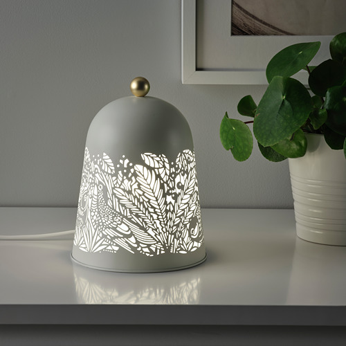 Una lámpara de mesa decorativa, SOLSKUR, con un diseño inspirado en la naturaleza, proporciona una iluminación ambiental suave, colocada sobre una superficie blanca junto a una planta en maceta.