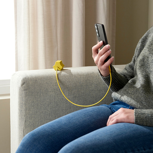 Persona en el sofá usando teléfono, organizador de cable HAVSKÅL, atuendo informal, cortinas detrás.