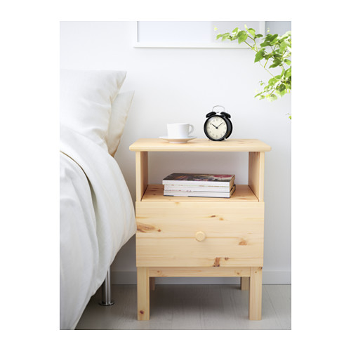 Una mesita de noche minimalista de madera clara junto a una cama, con libros y un tradicional despertador negro.