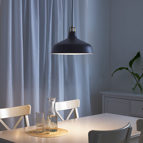 Comedor moderno con lámpara colgante negra y mesa blanca, iluminación focalizada en jarra y vasos, cortinas azules y sillas blancas.