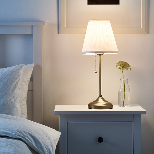 Una pequeña y elegante lámpara de noche ÅRSTID con pantalla blanca y base de acabado latón sobre una cajonera azul junto a una cama.