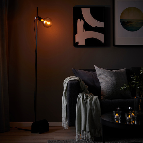 Habitación oscura con lámpara de pie negra HÅRSLINGA, sofá gris, mantas y arte en la pared.