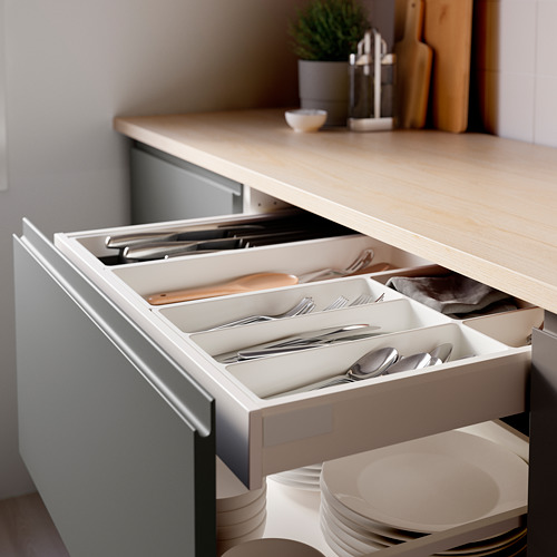 Cajón de cocina abierto con bandeja de utensilios organizada, que incluye tenedores, cucharas, cuchillos y espátula de madera. Encimera de madera de color claro visible arriba.