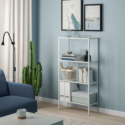Estantería moderna BAGGEBO de metal plateado con libros y jarrones contra la pared azul. Gran cactus y silla azul cerca.