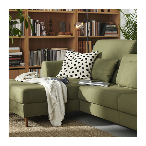 Moderno sofá en forma de L verde oliva con cojín punteado, patas de madera y ordenador portátil, junto a una estantería con artículos de decoración.