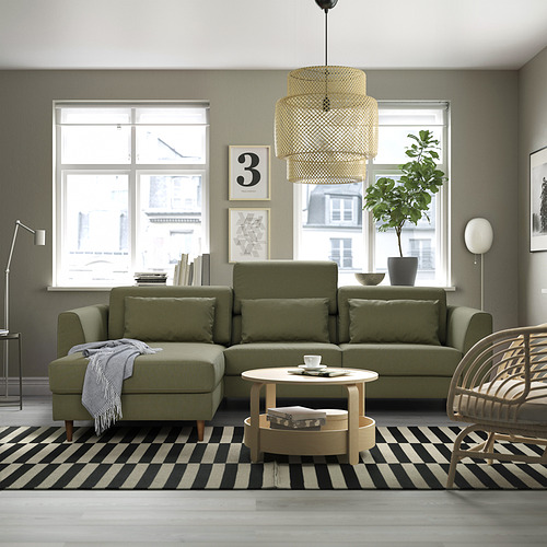 Salón moderno con sofá olivo, grandes ventanales, alfombra de rayas, mesa de centro, planta, silla de mimbre y luz escalonada.