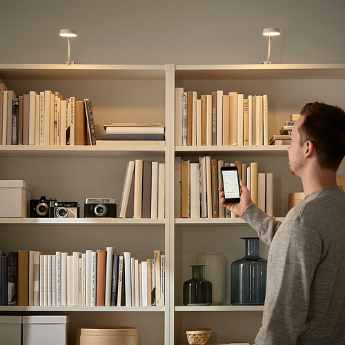 Un hombre ajusta la luz de un gabinete delgado y flexible unido a estantes llenos de libros y artículos, dirigiendo la luz con un brazo flexible.