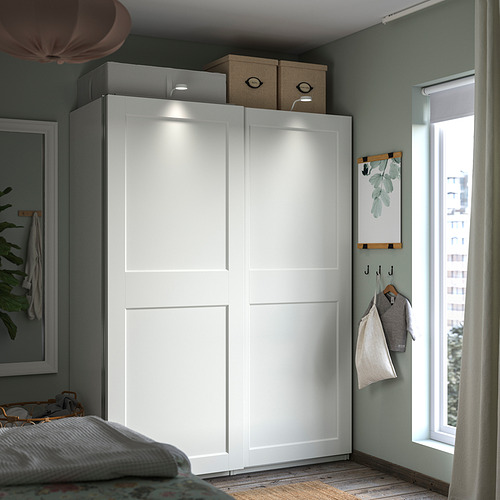 Moderno armario blanco PAX con puertas correderas, cajas de almacenamiento superiores, planta, perchas cerca de la ventana.