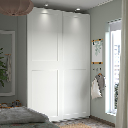 Gran armario blanco con puertas correderas en la esquina del dormitorio, junto a una ventana con cortinas, con perchas de madera y ropa gris.