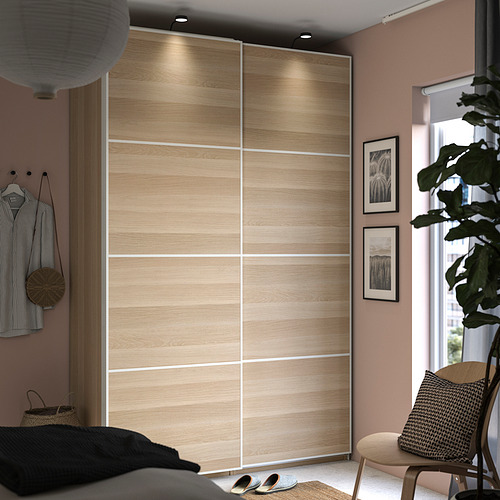 Dormitorio moderno con armario PAX, puertas correderas de madera clara, ropa tendida, planta grande, impresiones artísticas, silla acogedora.