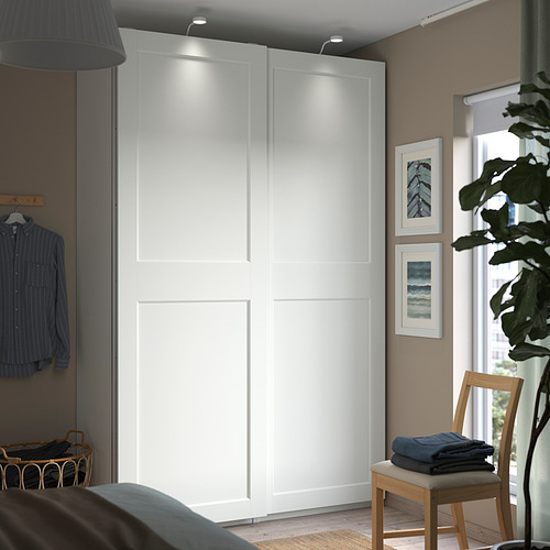 Dormitorio moderno con armario PAX, puertas correderas e iluminación. Ropa en perchero de pared, silla de madera, planta grande junto a la ventana. Estampados de la naturaleza en la pared.