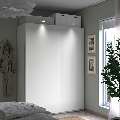 Dormitorio moderno con armario PAX blanco, puertas correderas, cortinas transparentes, paredes grises, plantas, prendas colgadas y obras de arte.