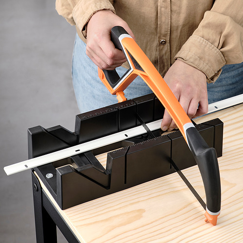 Persona cortando plástico con sierra de mango naranja sobre mesa de madera.