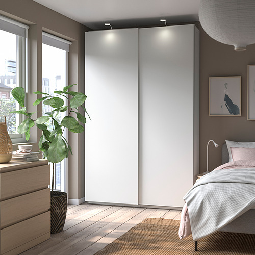 Dormitorio moderno con armario PAX blanco, suelo de madera, cama y planta grande.