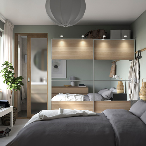 Elegante armario PAX en madera clara con puertas con espejos, iluminación tenue, estantes organizados y una acogedora cama con sábanas grises.